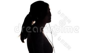 一个年轻快乐的女人戴着大耳机<strong>听</strong>音乐，拿着智能手机的<strong>剪影</strong>，女孩在白色的隔离巴上跳舞